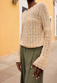 Pull en tricot beige avec un motif ajouré complexe, col rond et manches évasées, associé à un pantalon vert olive sur un trottoir.
