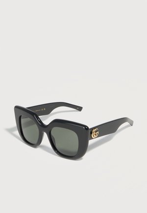 Lunettes de soleil carrées surdimensionnées noires avec des verres foncés et le logo double-G doré sur les branches, présentées sur une surface blanche.