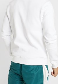 Sudadera blanca con cuello redondo y puños acanalados, que presenta una textura suave y un diseño ajustado, combinada con pantalones deportivos de color verde azulado.