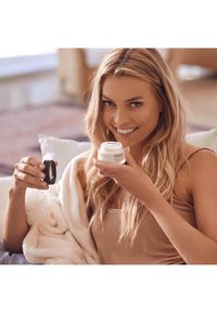 AHAAVA fuktighetskräm i en liten klar burk med ett vitt lock som hålls i ena handen; en mörk kompakt i den andra. Mjuk filt i bakgrunden.