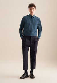 Teal button-up shirt gemaakt van gladde stof, gecombineerd met op maat gemaakte donkere broek. Het shirt heeft een zak; zwarte nette schoenen maken de outfit compleet.