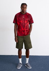Rød treningstrøye med svarte detaljer og geometrisk mønster, kombinert med grønne shorts, hvite sokker og grå sneakers.