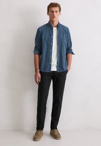 Blaues Hemd mit Knöpfen über einem weißen T-Shirt, schwarze Hose und beiger Schuhe. Das Modell steht vor einer hellen Wand, die Arme entspannt seitlich am Körper.