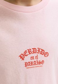 Šviesiai rožinė medvilninė marškinė su raudonais atspausdintais žodžiais: "PERDIDO en el PARAISO, BIENVENIDOS A PUERTO RICO" dekoratyviu šriftu. Apvalus iškirpimas.