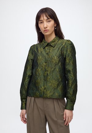 Femme aux longs cheveux foncés portant un chemisier vert à motifs floraux texturés et boutonné, ainsi qu'un pantalon plissé marron, debout devant un fond clair uni.