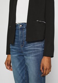 Černá zkrácená bunda s texturovaným povrchem, s přední kapsou na zip. Nositá na vysokém pasu modrých džínách s knoflíkovým zapínáním.