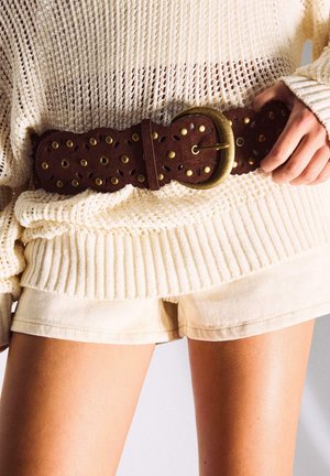 Person trägt einen cremefarbenen Strickpullover und Shorts, justiert einen breiten braunen Ledergürtel mit Messingnieten und großer ovaler Schnalle an der Taille.