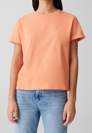 Femme portant un t-shirt uni pêche à manches courtes avec une petite broderie en forme de cœur, associé à un jean en denim bleu clair, sur un fond uni.