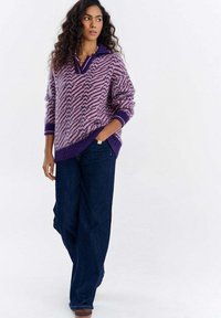 Kobieta o kręconych włosach, ubrana w fioletowy i różowy sweter w wzory oraz ciemnoniebieskie szerokie jeansy, stojąca z jedną ręką w kieszeni.