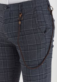 Pantalon à carreaux bleu marine avec poches latérales, fermeture par bouton, et une chaîne tressée en cuir marron attachée aux passants de ceinture.