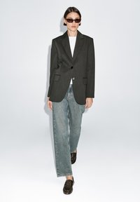 Donkergrijze op maat gemaakte blazer met gestructureerde schouders, twee voorzakken, over een witte T-shirt, gecombineerd met lichtblauwe rechtlopende jeans en zwarte instapschoenen.