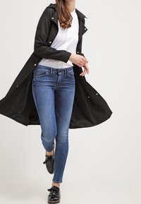 Svart trenchcoat med en strukturerad design, guldknappar och krage över en vit skjorta. Kombinationen med blå jeans och svarta skor.