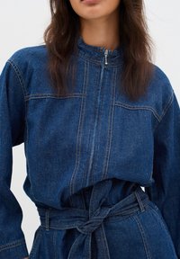 Dunkelblauer Denim-Overall mit Reißverschluss vorne, hohem Kragen, langen Ärmeln und einem gebundenen Gürtel in der Taille mit sichtbaren Nähdetails.