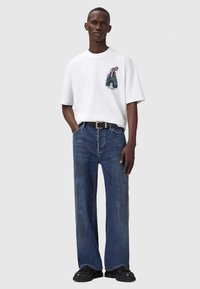 Muž v bílém grafickém tričku, modrých rovně střižených džínách, černém opasku a černých botách, stojí s jednou rukou v kapse.