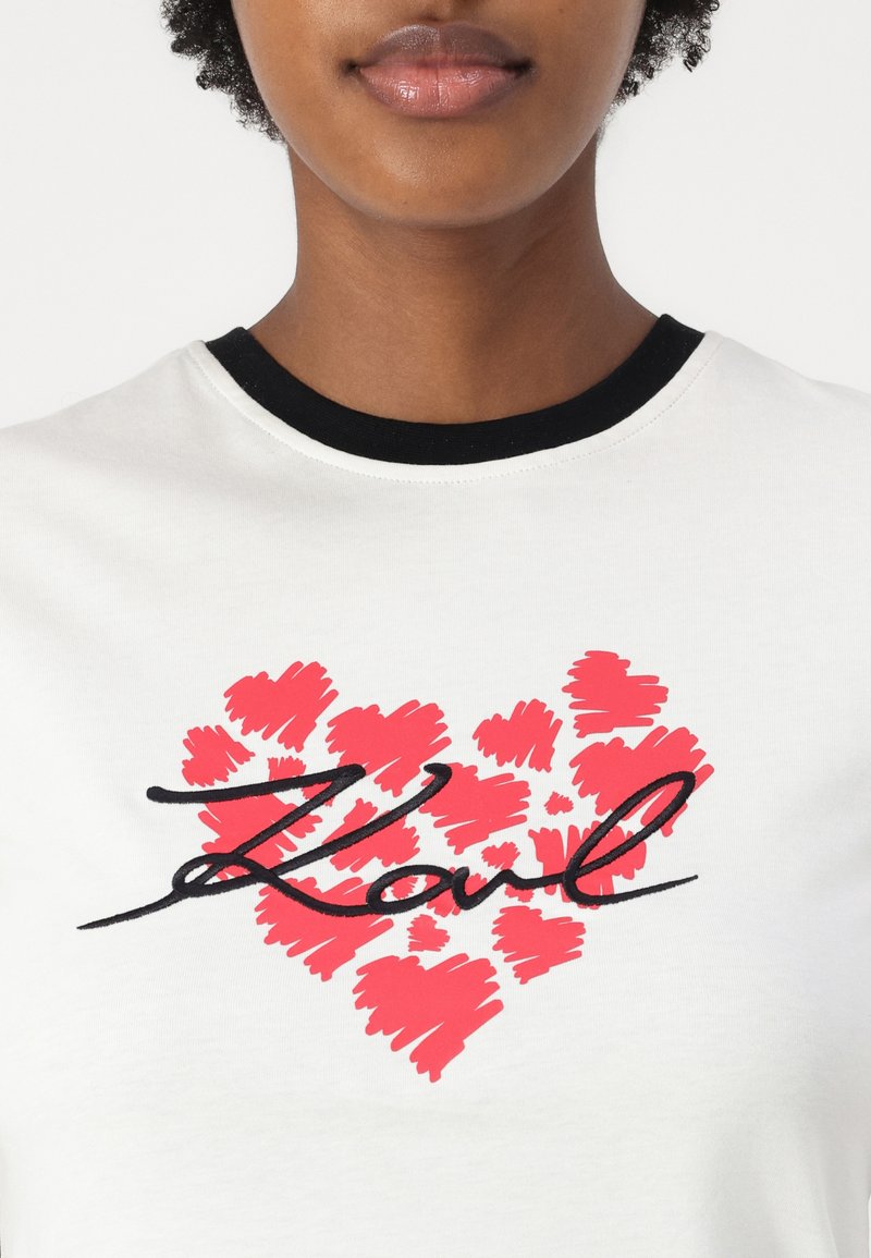 Weißes Baumwoll-T-Shirt mit einem schwarzen gerippten Rundhalsausschnitt und einem roten Herz-Graphic mit einer handgeschriebenen "Karl"-Unterschrift in Schwarz.