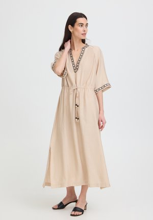Donna in piedi che indossa un lungo abito beige con rifinitura geometrica nera, una cintura in vita e sandali neri con tacco, guarda di lato con una mano vicino al collo.