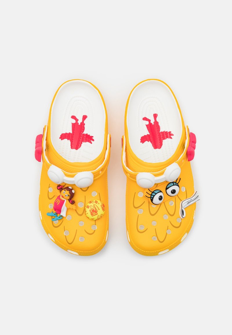 Sabots jaunes pour enfants avec des brides blanches, décorés d’yeux de dessin animé et d’un petit pendentif personnage sur un fond uni.
