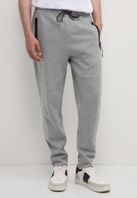Mann, der graue Jogginghosen mit schwarzen Reißverschlusstaschen und weiße Sneaker mit schwarzen und beigen Akzenten trägt, steht auf einem hellen Boden.