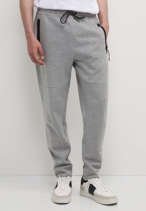 Mann, der graue Jogginghosen mit schwarzen Reißverschlusstaschen und weiße Sneaker mit schwarzen und beigen Akzenten trägt, steht auf einem hellen Boden.