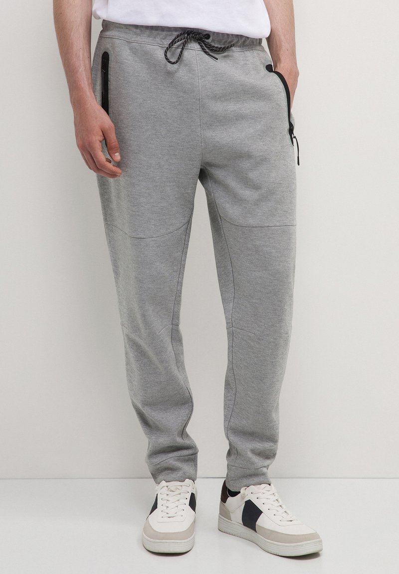 Mann, der graue Jogginghosen mit schwarzen Reißverschlusstaschen und weiße Sneaker mit schwarzen und beigen Akzenten trägt, steht auf einem hellen Boden.