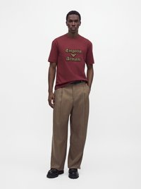 Мъж, носещ червена тениска Emporio Armani, кафяв широк панталон и черни обувки, стои с една ръка в джоба пред бял фон.