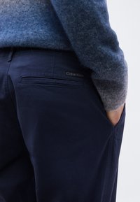 Άτομο που φοράει μπλε σκούρο παντελόνι Calvin Klein με το δεξί χέρι στην τσέπη και γαλάζιο-γκρι πουλόβερ με γυρισμένη μανσέτα.