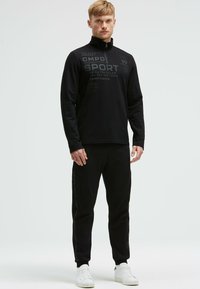 Schwarzer Sportpullover mit einem Reißverschlusskragen und bedruckten Textdetails. Kombiniert mit schwarzen Jogginghosen mit seitlichen Logoakzenten und weißen Sneakers.