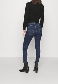Jeans de mezclilla oscuros ajustados con costuras en contraste y dos bolsillos traseros. Combinados con botas negras hasta el tobillo y un suéter negro texturizado.