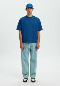 Blå bomulds t-shirt med boxy pasform, parret med lyseblå jeans. Svarte og hvite sko, med en blå kasket med hvid tekst.