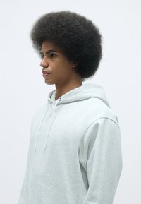 Felpa con cappuccio di un grigio chiaro, dal tessuto morbido e dotata di lacci regolabili, indossata da una persona con un'acconciatura afro voluminosa.