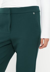 Tamnozeleni krojeni hlače na osobi koja nosi bijelu majicu dugih rukava, s otkrivenim strukom, detaljima džepa i dijelom ruke.