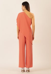 Jumpsuit coral de un solo hombro con drapeado asimétrico, diseño de pierna ancha y textura suave. Combinado con zapatos de tacón cuadrado plateados.