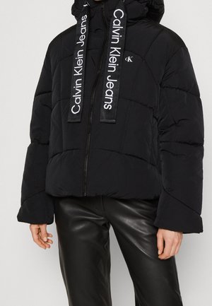 Zwarte donzen jas met grote capuchon, rits, brede mouwen en "Calvin Klein Jeans" gedrukt op de hangende trekkoorden, gecombineerd met zwarte leren broeken.