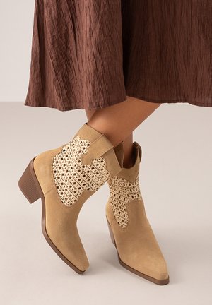 Botas ankle de ante beige con un detalle de crochet y punta afilada; tacón apilado y parte superior texturizada. Combinadas con una falda marrón plisada.
