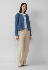 Denimjas met lange mouwen, borstzakken en knoopsluiting, gedragen over een lichtblauw gestreept shirt en beige wijde broek.