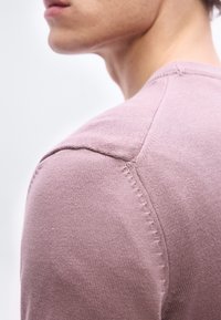 Κοντινό πλάνο στον ώ μο και τον λαιμό ενός ατόμου που φοράει ανοιχτό μοβ πλεκτό πουλόβερ, με κατεύθυνση προς τα αριστερά, μπροστά σε απλό λευκό φόντο.