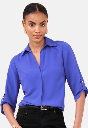 Blaue Bluse zum Knöpfen mit V-Ausschnitt und aufgekrempelten Ärmeln. Aus glattem Stoff. Kombiniert mit schwarzer Hose und schwarzem Gürtel.