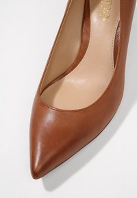 Brun läder högklackad sko med spetsig tå och slät yta. Har en lågskuren design på sidorna och neutral innervägg.