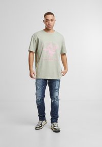 Ljusgrön oversized t-shirt med rosa tryck som har vingar och text. Bärs med slitna mörkblå jeans och svartvita sneakers.