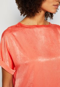 Blouse en satin corail avec une coupe décontractée, dotée d'un large col et de manches retroussées, texture lisse et éclat discret.
