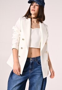 Blazer bianco doppiopetto con bottoni dorati, abbinato a un top bianco corto e jeans a gamba larga in denim scuro a vita alta.