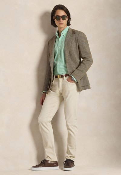 Γκρι σακάκι herringbone πάνω από ανοιχτό πράσινο πουκάμισο, εκρού παντελόνι και καφέ αθλητικά παπούτσια. Αξεσουάρ περιλαμβάνουν γυαλιά ηλίου και ταμπά ζώνη.