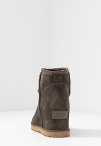 Bota UGG de ante marrón con suela de goma color tan, costuras visibles y etiqueta trasera sobre un fondo blanco, en ángulo hacia una esquina.