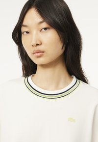 Felpa bianca con colletto a coste e strisce nere, bianche e giallo neon. Logo Lacoste in verde chiaro ben visibile sul petto.