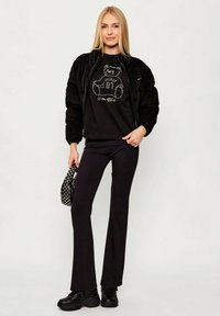 Czarna kurtka bomberka z teksturowanej tkaniny, czarny t-shirt z grafiką niedźwiedzia, czarne rozkloszowane spodnie oraz czarne sneakersy.