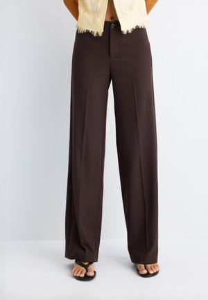 Femme portant un pantalon large brun foncé taille haute avec pli sur le devant, associé à des sandales noires en tongs, sur un fond clair uni.