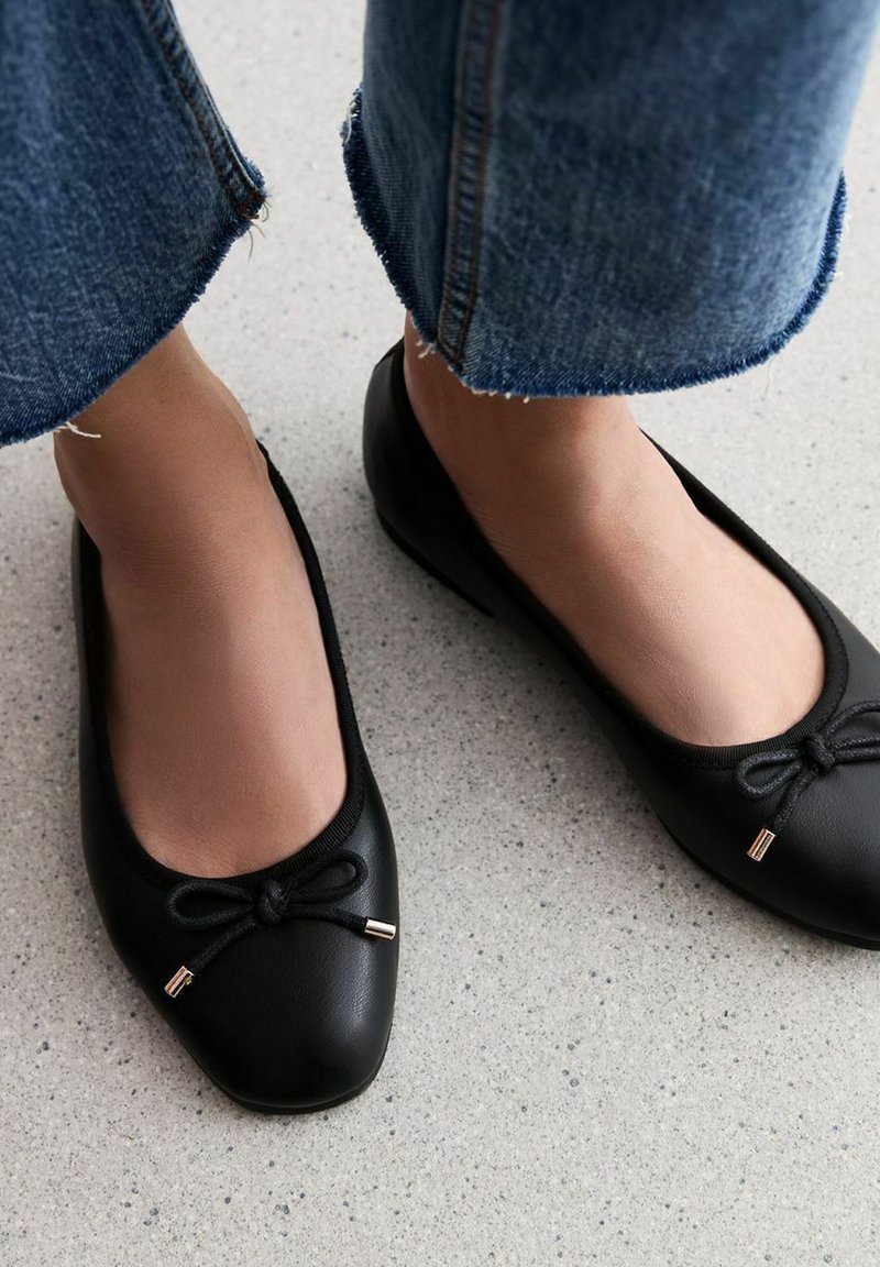 Ballerines en cuir noir avec un bout rond, ornées d'un nœud décoratif et d'un accent doré. Portées avec un jean en denim retroussé.