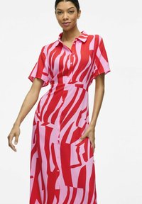 Vestido de patrones rojos y rosas con mangas cortas, cuello y cierre frontal de botones. La tela parece suave y ligera.