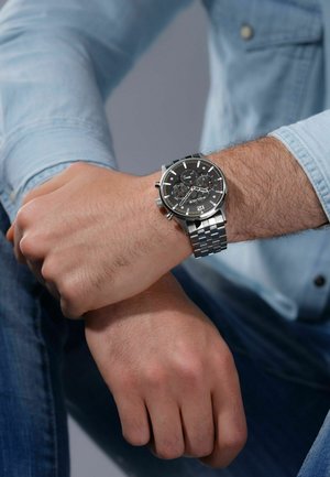 Orologio in acciaio inox argentato con quadrante nero, funzionalità cronografo e bracciale a maglie. Il polso è parzialmente coperto da una camicia di jeans.