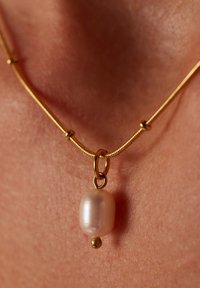Collier en or présentant un pendentif en perle ovale lisse avec un petit accent de boule en or en bas, posé sur une texture de peau.